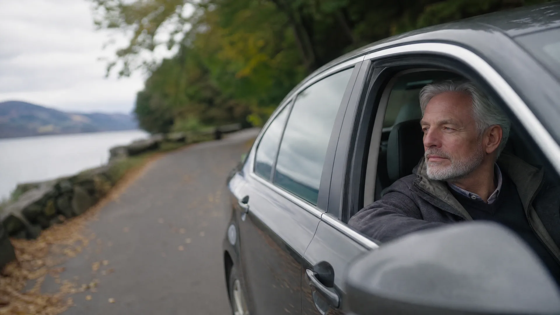 A grey-haired bearded man looks out of the open rear window of a dark sedan parked on a leaf-strewn road beside a lake, with wooded hills in the background under an overcast sky.