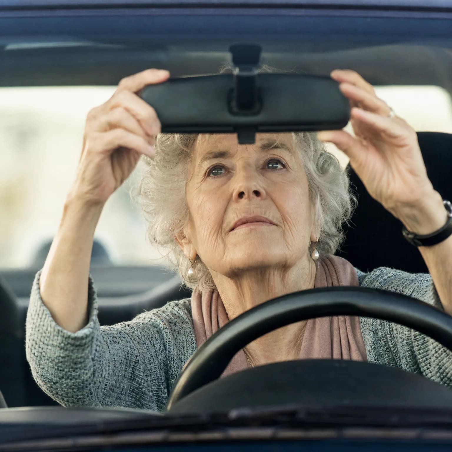 An older woman with short grey hair, seated behind a car steering wheel, reaches up with both hands to adjust the rearview mirror, seen through the windshield.