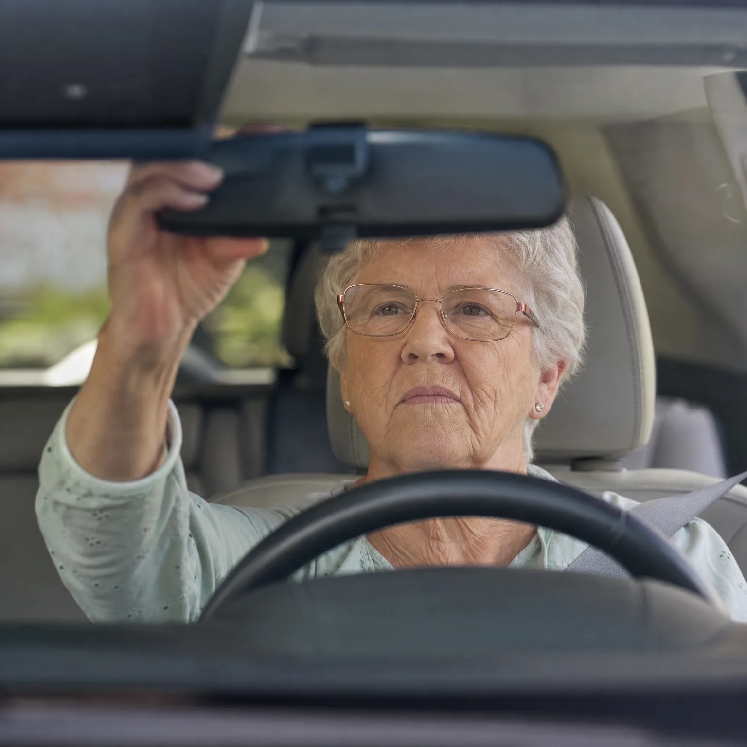 An older woman with short gray hair and glasses sits in a car's driver seat behind the steering wheel, reaching up with her left hand to adjust the rearview mirror, seen through the windshield.