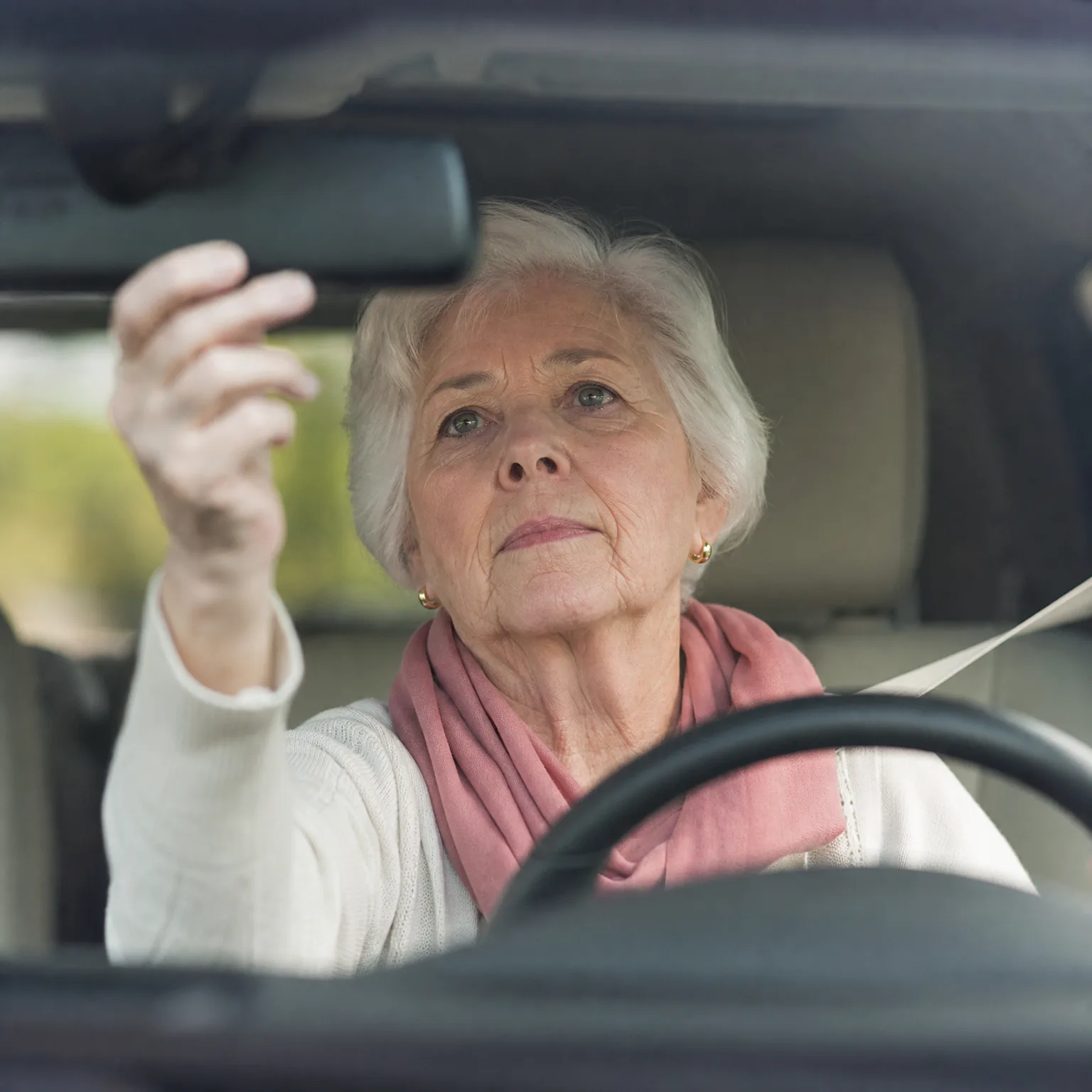 An older woman with short white hair, wearing a white top and pink scarf, sits in a car behind the steering wheel and reaches up to adjust the rearview mirror.