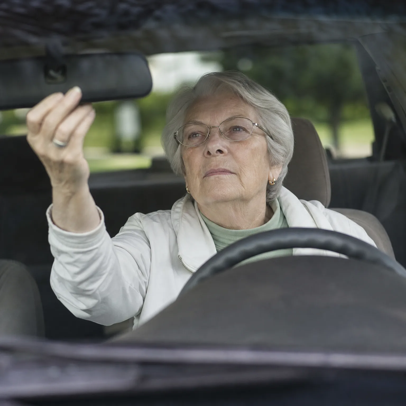An older woman with gray hair and glasses, wearing a white jacket, sits in a car's driver seat and reaches up to adjust the rearview mirror, photographed through the windshield with green trees blurred behind.