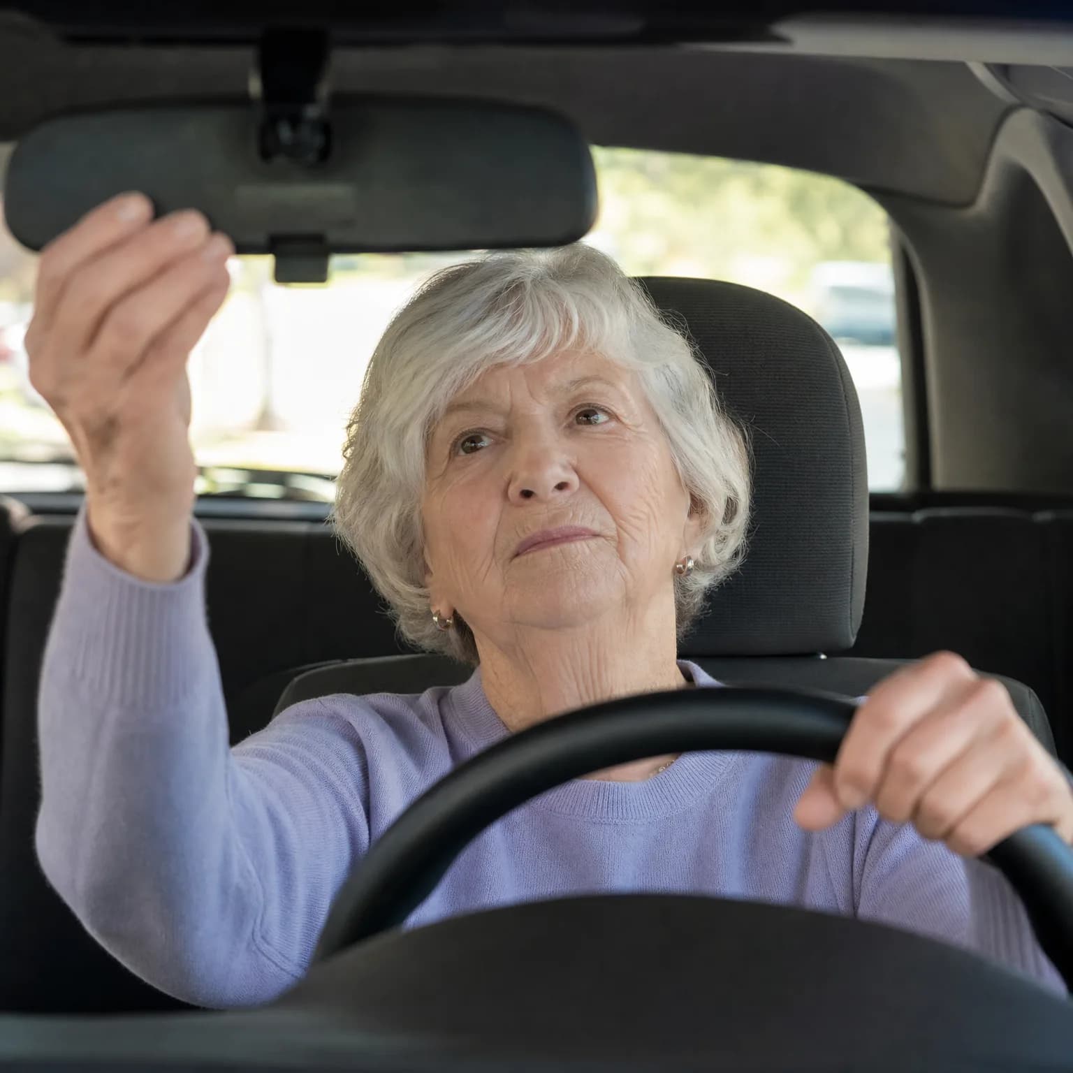 An older woman with short white hair in a light purple sweater sits in a car's driver seat, one hand on the steering wheel and the other adjusting the rear-view mirror.
