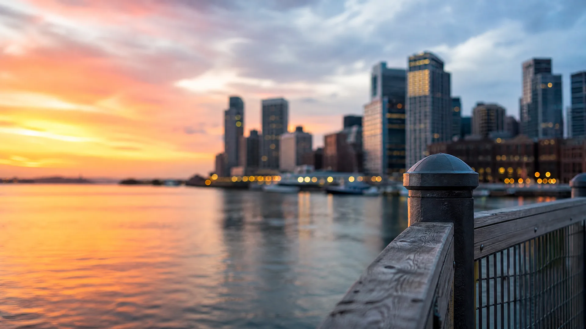 A wooden railing with a capped post in sharp focus in the foreground, with calm water and a blurred city skyline of high-rise buildings under an orange and blue sunset sky behind.
