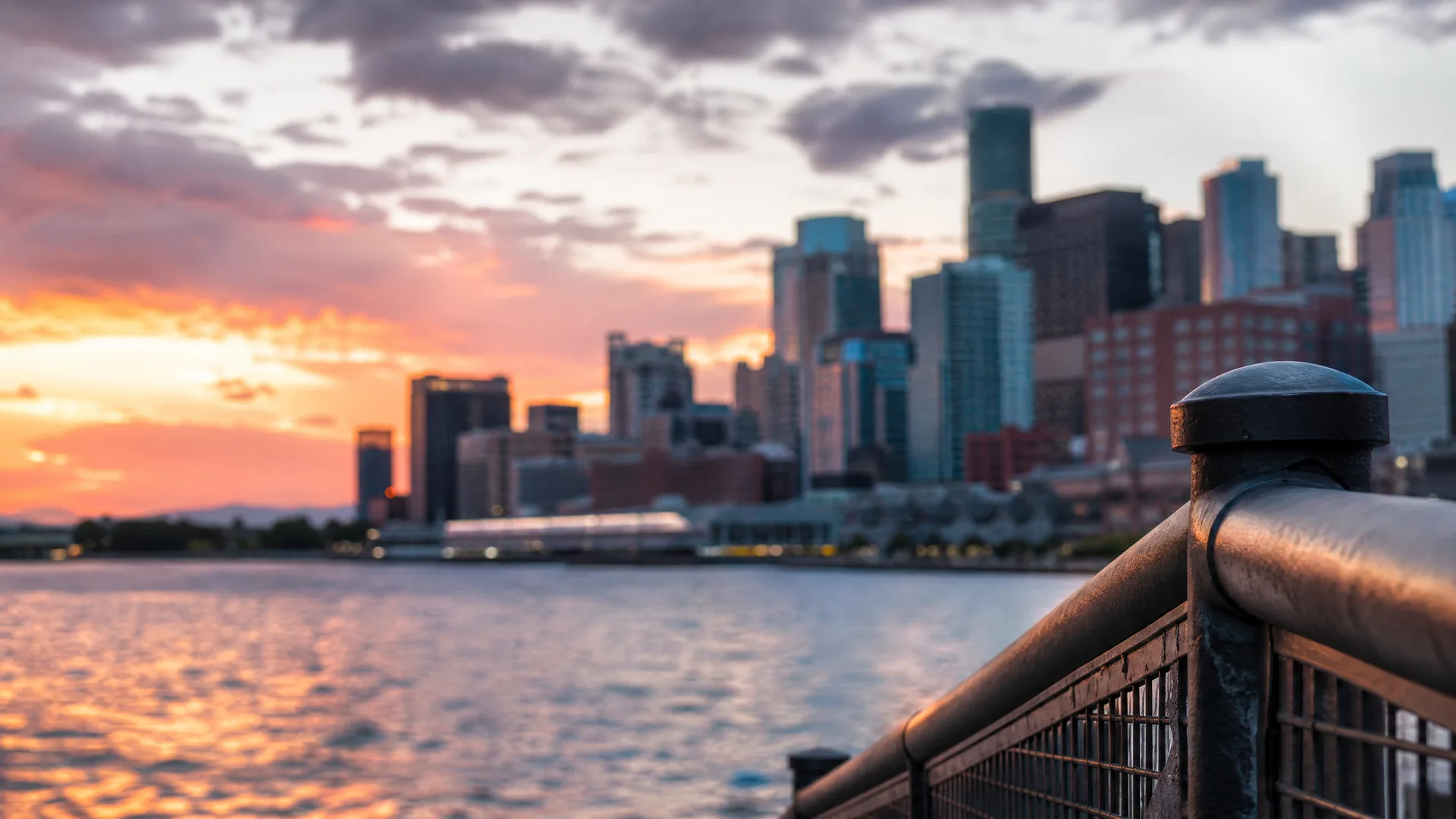 A city skyline of high-rise towers across a body of water at sunset, with orange clouds and a blurred metal railing in the right foreground.