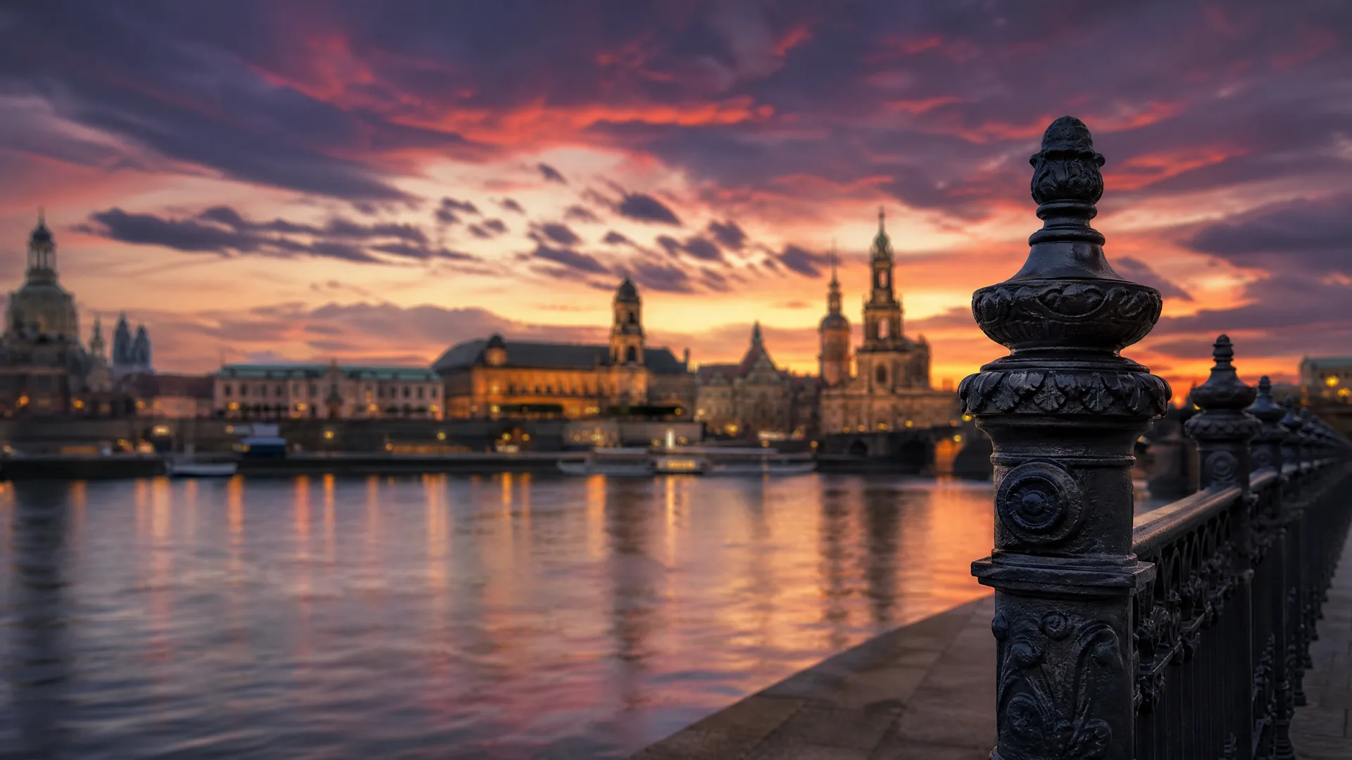 A river at sunset with an illuminated city skyline of domed and towered buildings on the far bank, seen past an ornate dark metal railing post in the foreground.